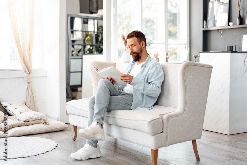 Young bearded handsome man reading book on couch at home Young bearded handsome man reading book on couch at home