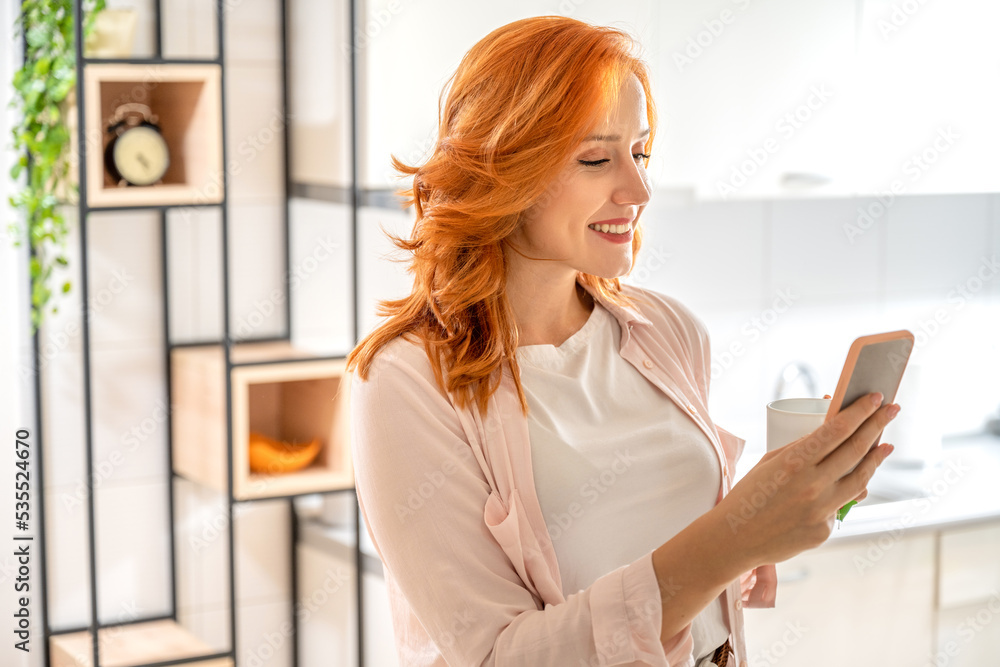 Portrait of young ginger woman drinking tea while using a phone and standing in kitchen at home in the morning Portrait of young ginger woman drinking tea while using a phone and standing in kitchen at home in the morning