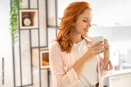 Beautiful smiling ginger girl standing, wearing casual clothes, drinking tea and relaxing at home in the morning