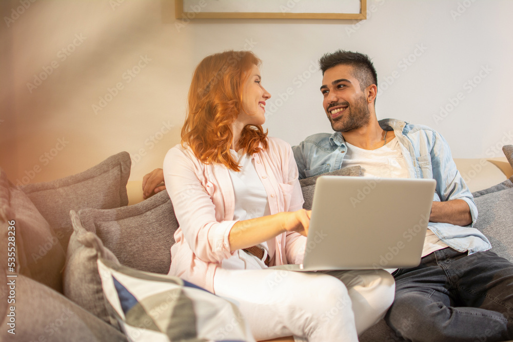 Cute happy couple looking to each other and using laptop while sitting on sofa at home. Cute happy couple looking to each other and using laptop while sitting on sofa at home.