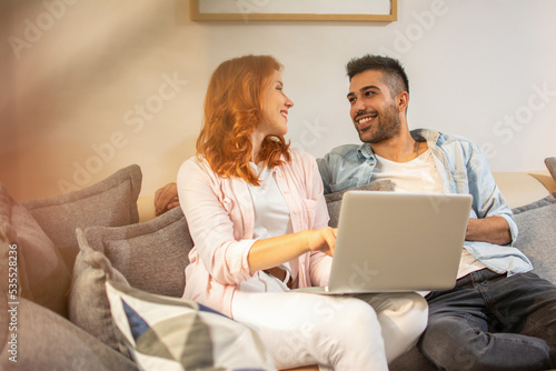 Cute happy couple looking to each other and using laptop while sitting on sofa at home.