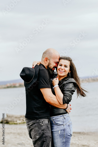 Portrait of happy    smiling  Couple standing on cliff against sea during  the winter .Young happy Bearded muscular  man  ikissing and hugging beautiful woman  in leather jacket and hat on a beach © Striker777