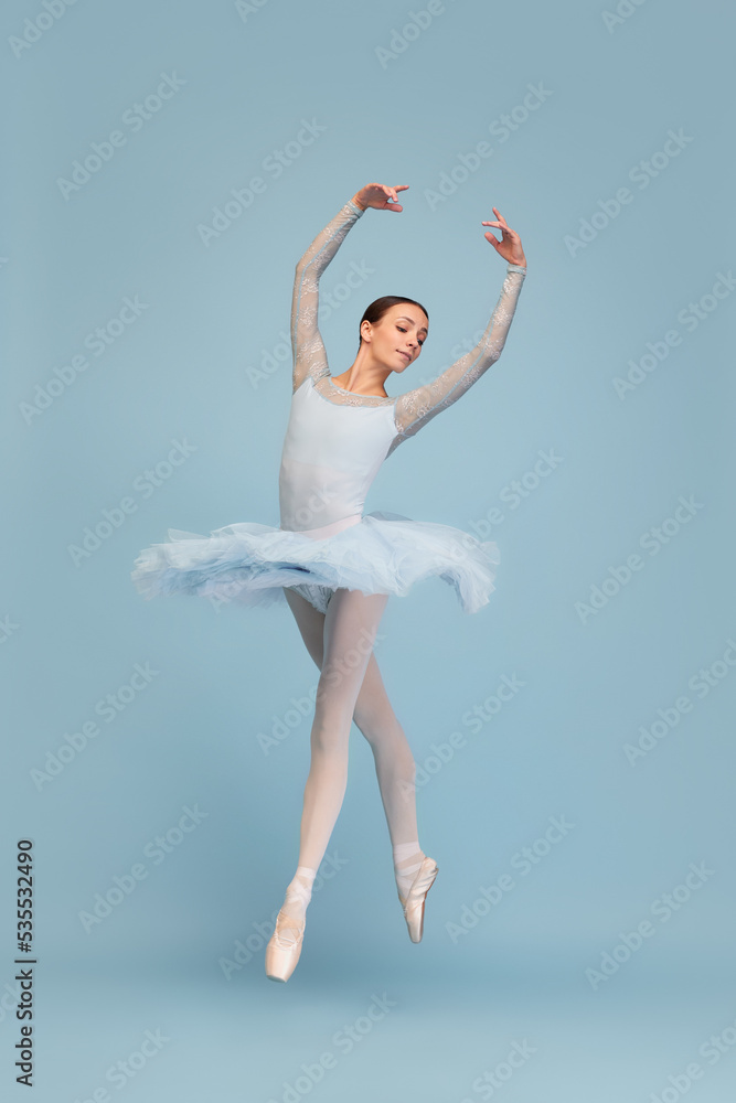Portrait of tender young ballerina dancing, performing isolated over blue studio background. Beauty of choreography