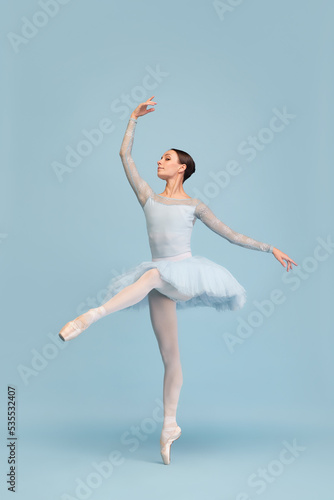 Portrait of tender young ballerina dancing, performing isolated over blue studio background. Graceful theatrical performance