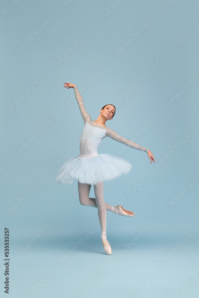 Portrait of tender young ballerina dancing, performing, posing in movement isolated over blue studio background