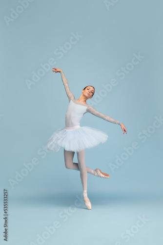 Portrait of tender young ballerina dancing, performing, posing in movement isolated over blue studio background