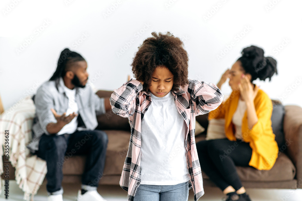 Obraz premium Unhappy sad little african american girl, closed her eyes and ears, feel lonely lacking parents attention, suffer from domestic family conflicts, parents on the background shouting at each other