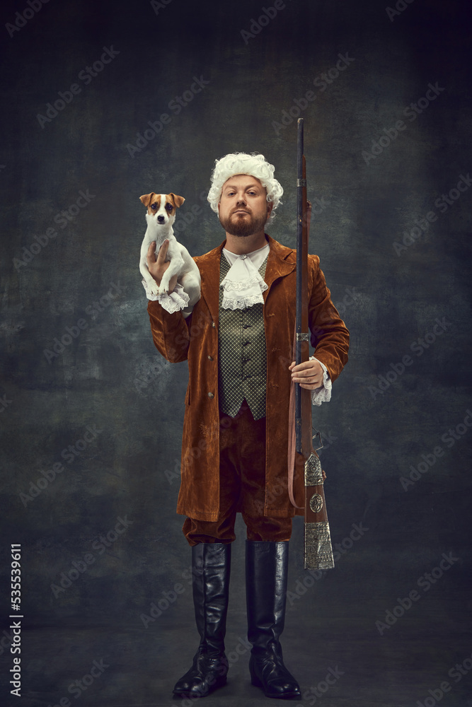 Hunter with doggy. Portrait of young man in image of nobleman in vintage brown hunting suit and white wig with old hunting rifle isolated over dark retro background.
