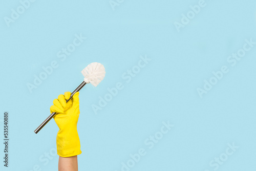 Mano con guante de goma amarillo sosteniendo una escobilla de limpieza sobre un fondo celeste pastel liso y aislado. Vista de frente y de cerca. Copy space