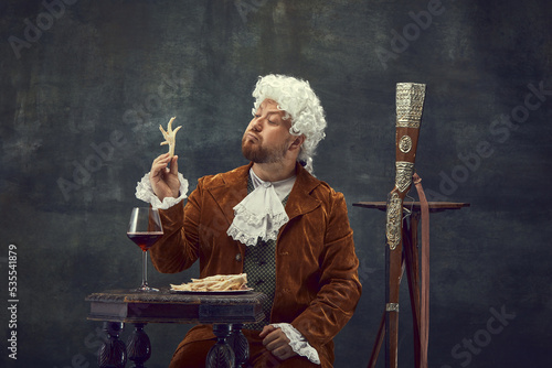 Strange things. Vintage portrait of young man in brown vintage suit and white wig like medieval royal hunter isolated on dark background.
