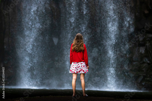 A girl in a red sweatshirt and red and white skirt stands over a powerful tropical waterfall in australia  millaa millaa falls in queensland  queensland rainforest