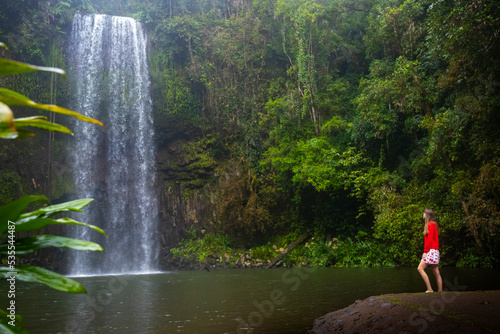 A girl in a red sweatshirt and red and white skirt stands over a powerful tropical waterfall in australia; millaa millaa falls in queensland; queensland rainforest photo