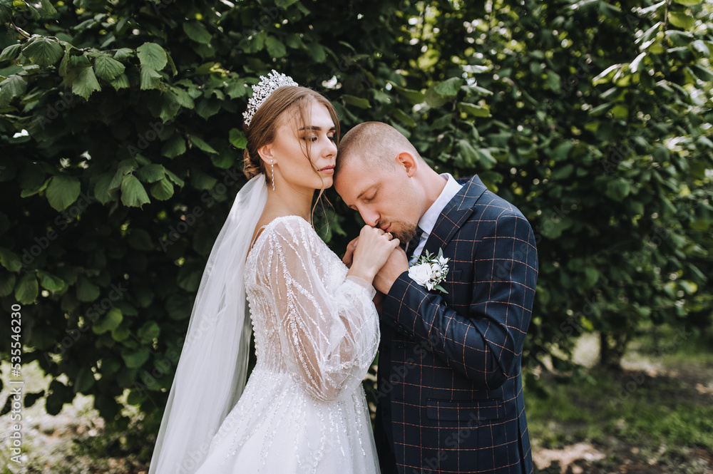 A young groom in a blue plaid suit and a beautiful bride in a white lace dress with a diadem, a bouquet in her hand gently hug, kiss in a park in nature. Wedding photography of the newlyweds.