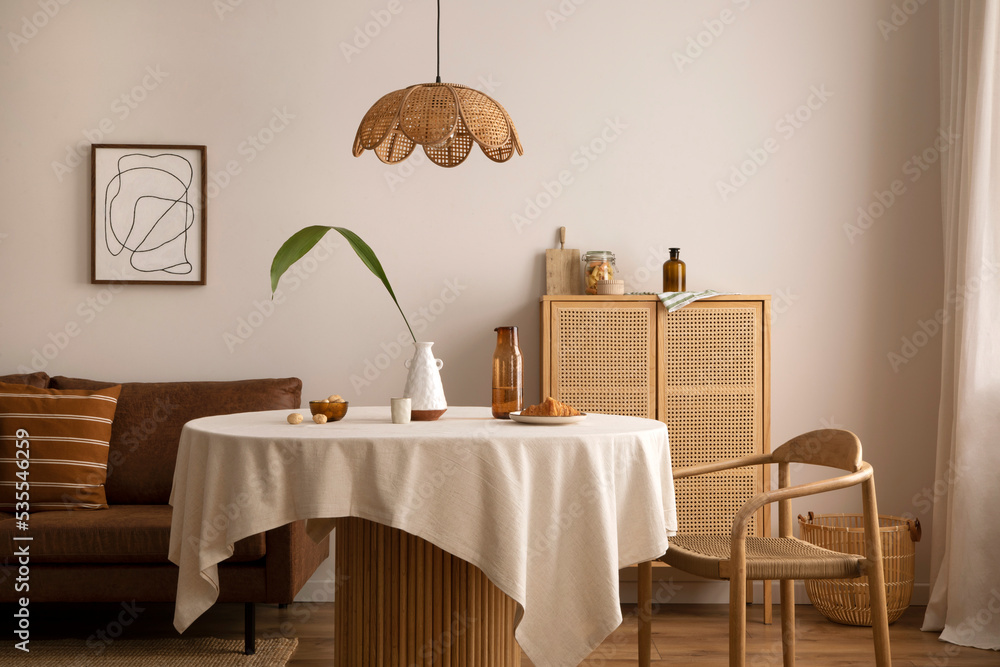 Fotka „Boho and cozy interior of dining room with round wooden table,  design chair, brown sofa, commode, mock up poster frame, pendant lamp, tea  pot, decoration and elegant accessories. Stylish home decor.“ ze