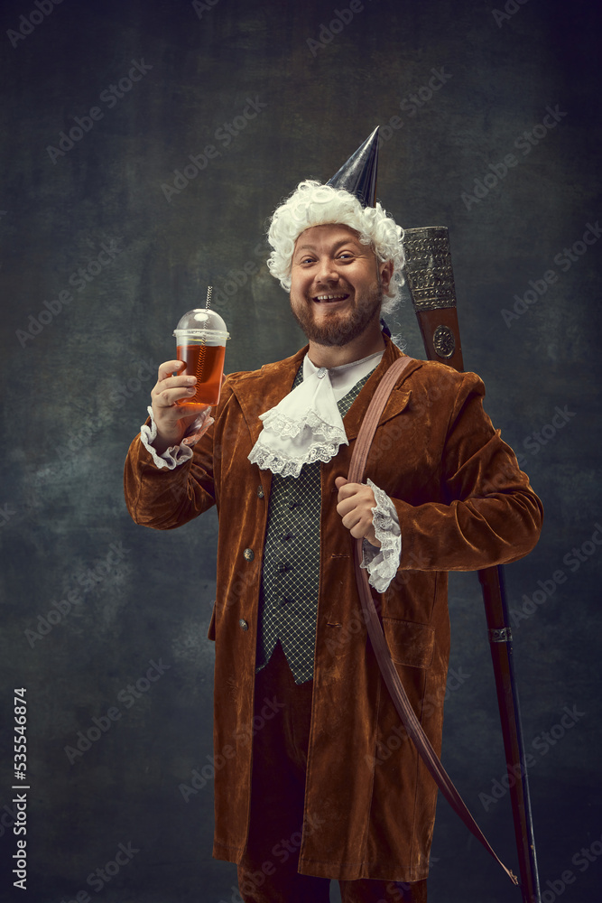 Beer party. Happy young man in brown vintage suit and white wig like medieval royal hunter isolated on dark background.
