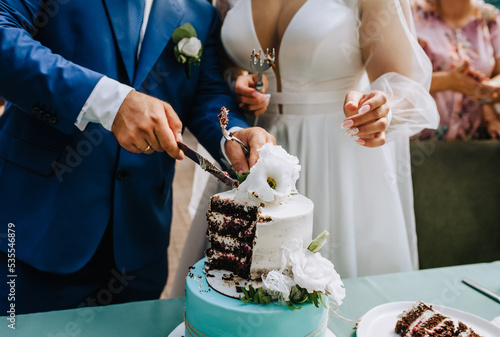 The bride and groom cut the sweet, delicious wedding cake on the table with a knife into large pieces on a plate at the ceremony. Photography, dessert.