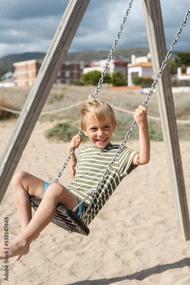 A happy boy on a swing on a sandy beach against the backdrop of apartment buildings and a stormy sky