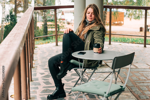 Young woman sitting on outdoor terrace in the forest drinking a cup of hot coffee. Beautiful female relax and enjoy outdoor activity lifestyle in nature on autumn travel vacation © MarijaBazarova
