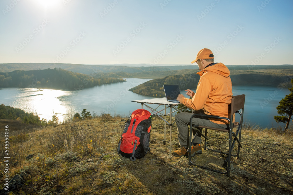 Fototapeta premium Man working on laptop on top mountains and relaxes near river at sunset. Freedom, remote work, freelancer, technology, internet, travel and vacation concepts.