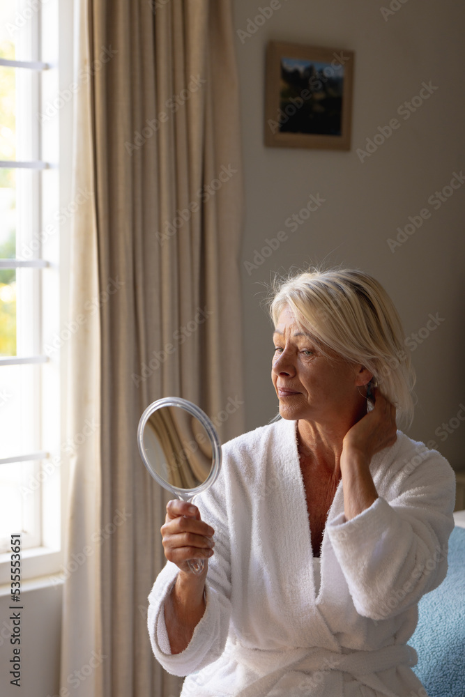Happy senior caucasian woman sitting on bed in bedroom, looking at mirror Happy senior caucasian woman sitting on bed in bedroom, looking at mirror
