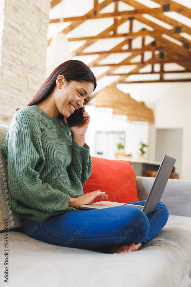 Happy biracial woman sitting on sofa in living room, using laptop and talking on smartphone Happy biracial woman sitting on sofa in living room, using laptop and talking on smartphone