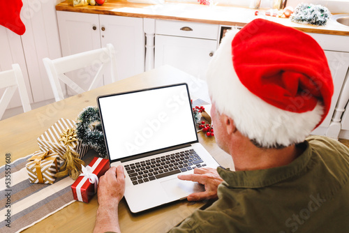 Caucasian man wearing santa hat, sitting at table in kitchen, using laptop with copyspace © wavebreak3