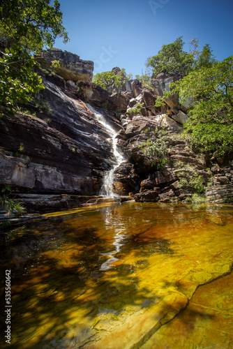 Small waterfall in the middle of the forest with yellowish water in the foreground and blue sky in the background