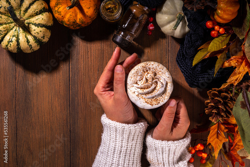 Girls hands with glass cup with tasty pumpkin spice latte, with white and orange pumpkin squash, pumpkin pie spices and autumn decor copy space. Traditiional autumn Thanksgiving holiday drink photo