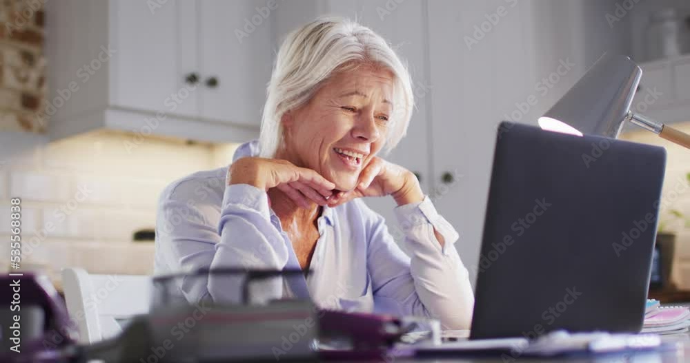 Happy senior caucasian woman sitting at table in kitchen, using laptop for online meeting Happy senior caucasian woman sitting at table in kitchen, using laptop for online meeting