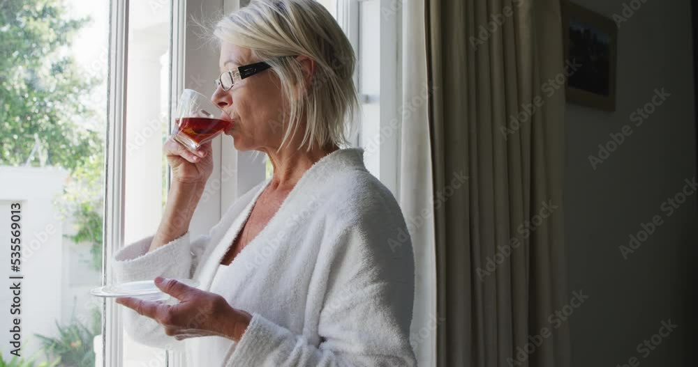 Happy senior caucasian woman standing in bedroom, looking through window and drinking tea Happy senior caucasian woman standing in bedroom, looking through window and drinking tea