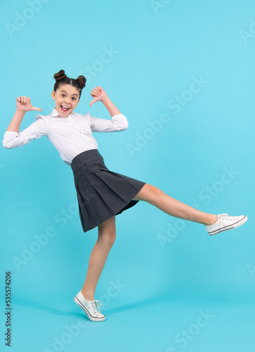 Excited schoolgirl, positive and smiling emotions of teen girl. Full length jump of teenager girl on blue isolated studio wear casual skirt and shirt. Funny school girl, amazed kid genius.