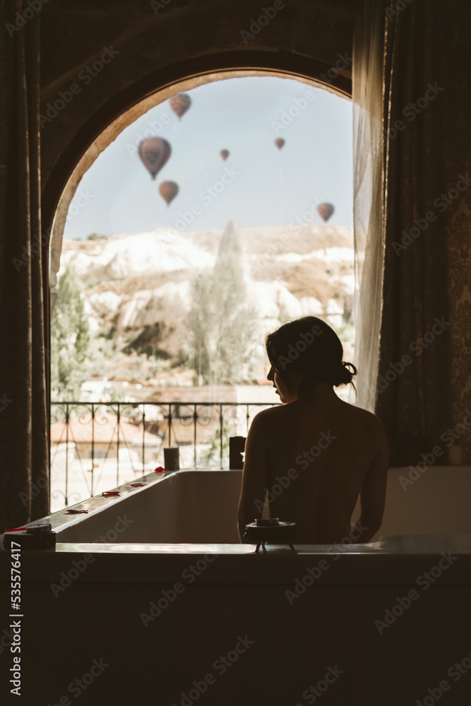 Silhouette of an unrecognizable young woman taking a bath in front of a window with hot air balloons flying in the background in the landscape - Cappadocia, Turkey