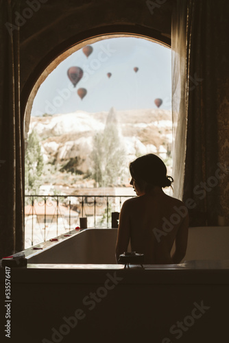 Silhouette of an unrecognizable young woman taking a bath in front of a window with hot air balloons flying in the background in the landscape - Cappadocia  Turkey