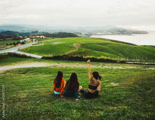 three women sitting on the grass against sea landscape with sunset