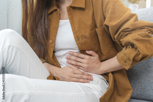Flatulence asian young woman hand in stomach ache, suffer from food poisoning, abdominal pain and colon problem, gastritis or diarrhoea sitting on sofa at home. Patient belly, abdomen or inflammation.