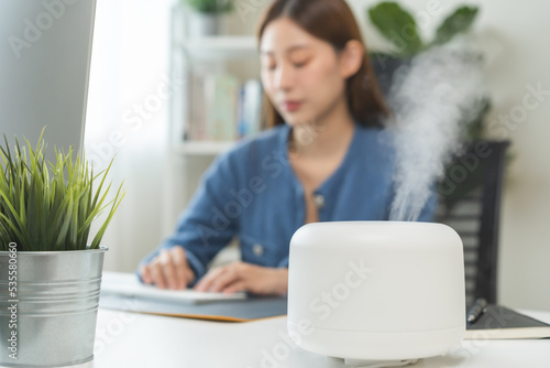 Tableau sur toile Modern air humidifier device during sitting workplace, happy asian young woman using computer work on internet, enjoying aromatherapy steam scent from essential oil diffuser in room at home office