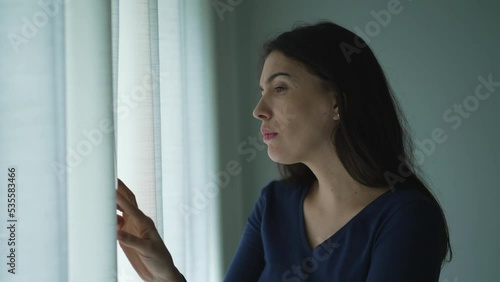 Anxious Woman standing by window peeking out in suspicion feeling distrust