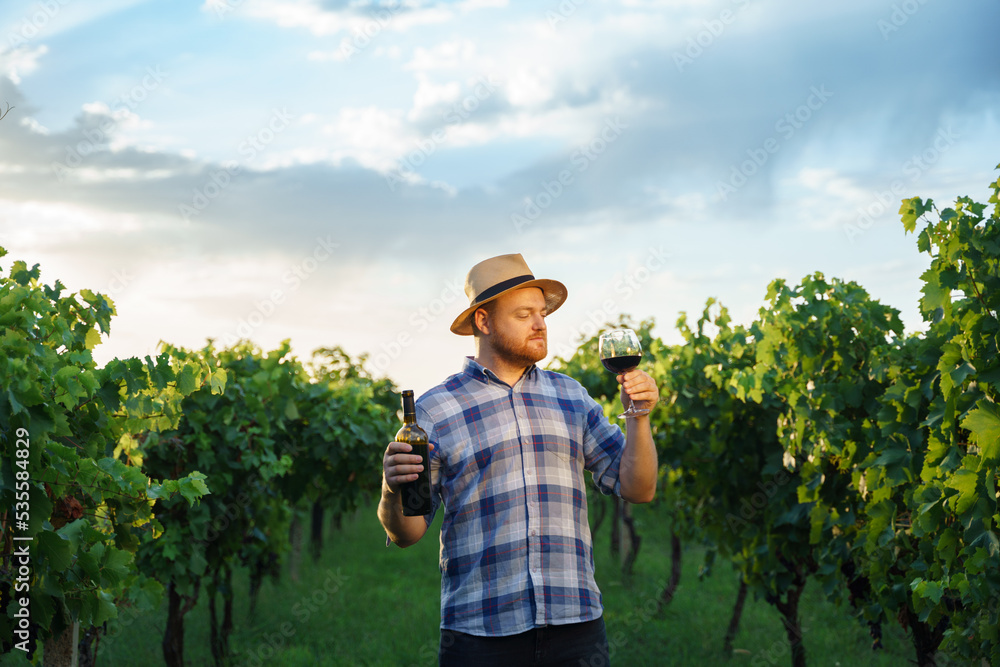 Portrait of a young, millennial vintner holding a glass and a bottle of organic bio red wine outdoors in a vineyard - Vine-growing, and wine-tasting concept in a rural winery