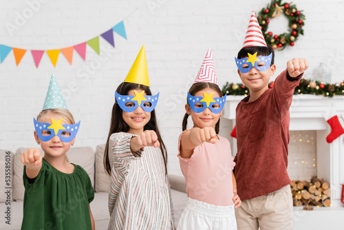 Cheerful interracial kids in party caps and masks gesturing during birthday party at home