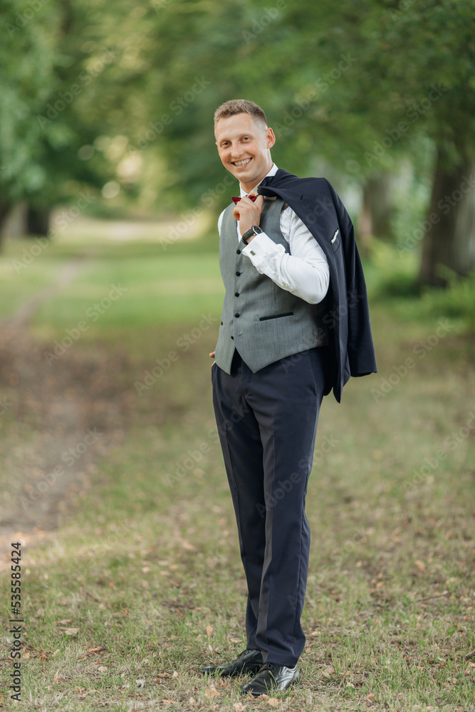 Portrait of young smiling man groom with short hair, wear blue suit, grey vest, red bow tie, standing in park in summer. Portrait of young smiling man groom with short hair, wear blue suit, grey vest, red bow tie, standing in park in summer.