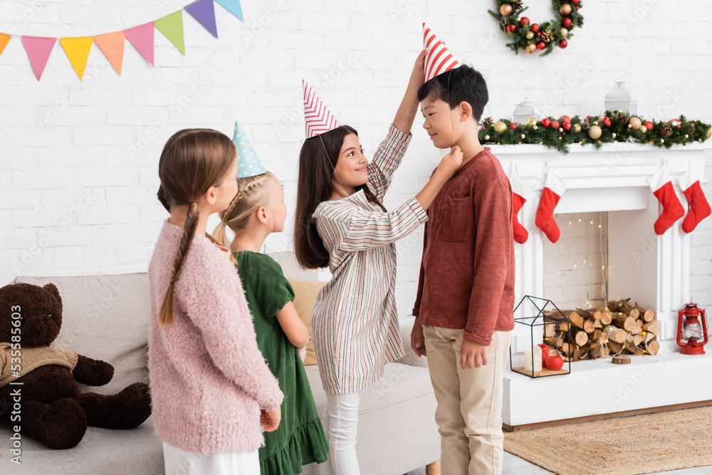 Obraz premium Smiling girl wearing party cap on asian friend during birthday party and christmas celebration