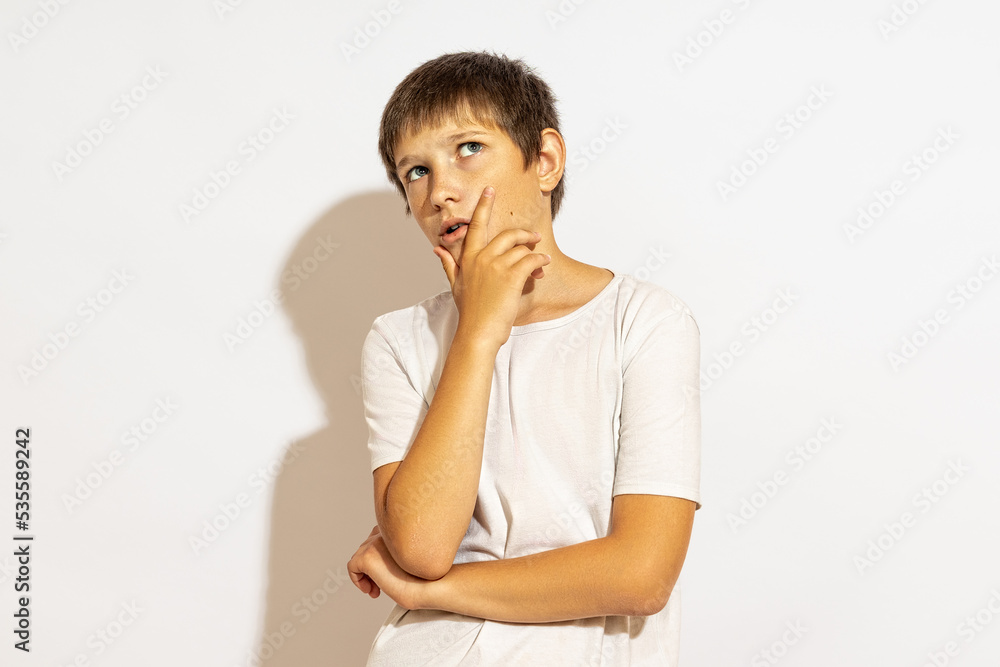 The teenager looks up thoughtfully. He put his palm on his chin. Hard light, white background.