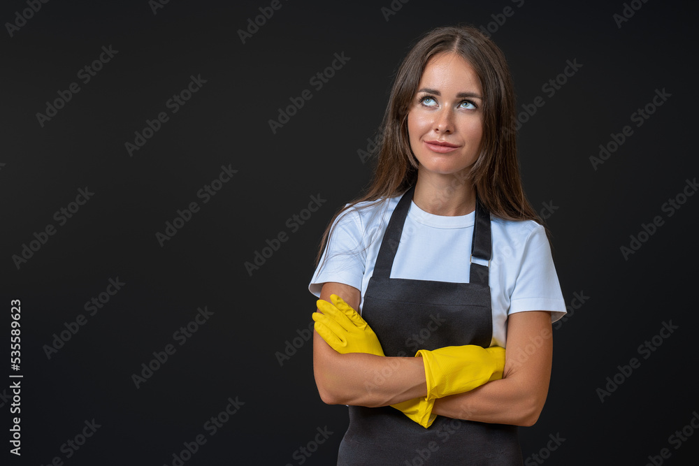 a housewife in an apron and gloves with flowing hair crossed her arms over her chest and thinks, stands on a black background with space for text a housewife in an apron and gloves with flowing hair crossed her arms over her chest and thinks, stands on a black background with space for text