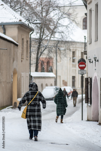 Beautiful narrow street in Vilnius Old Town with people walking in the storm, old buildings and street lamps in winter covered by the snow, vertical
