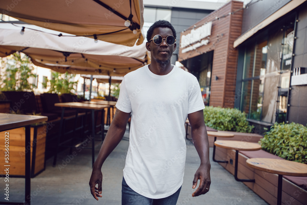 Fototapeta premium An african american man in a white t-shirt stands in a street cafe. Mock-up.