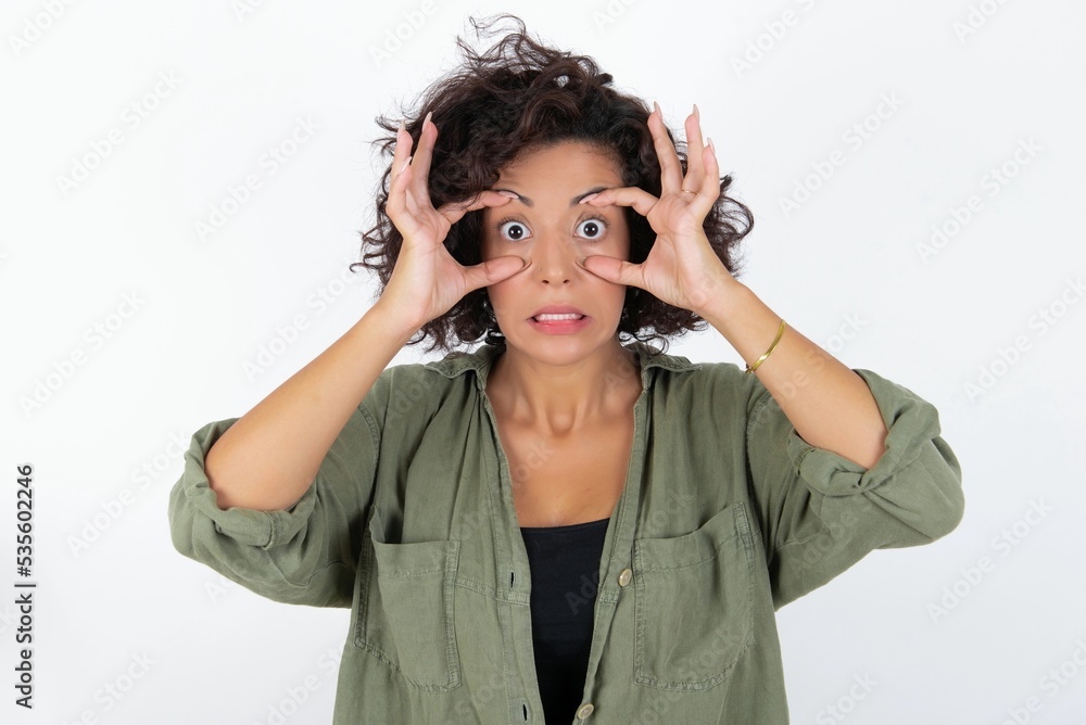 young beautiful woman with curly short hair wearing green overshirt over white wall keeping eyes opened to find a success opportunity. young beautiful woman with curly short hair wearing green overshirt over white wall keeping eyes opened to find a success opportunity.