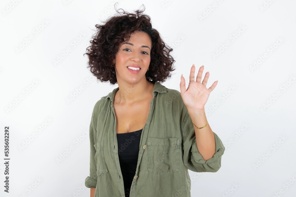 young beautiful woman with curly short hair wearing green overshirt over white wall Waiving saying hello happy and smiling, friendly welcome gesture. young beautiful woman with curly short hair wearing green overshirt over white wall Waiving saying hello happy and smiling, friendly welcome gesture.