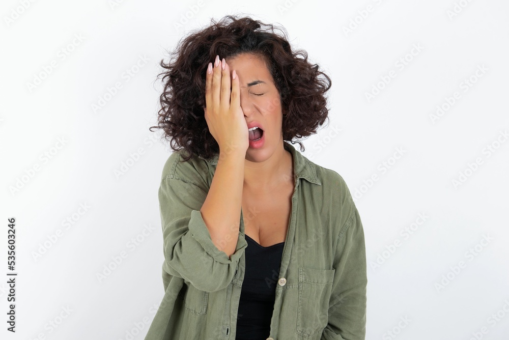 young beautiful woman with curly short hair wearing green overshirt over white wall Yawning tired covering half face, eye and mouth with hand. Face hurts in pain. young beautiful woman with curly short hair wearing green overshirt over white wall Yawning tired covering half face, eye and mouth with hand. Face hurts in pain.