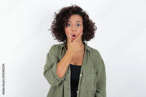 young beautiful woman with curly short hair wearing green overshirt over white wall Looking fascinated with disbelief, surprise and amazed expression with hands on chin