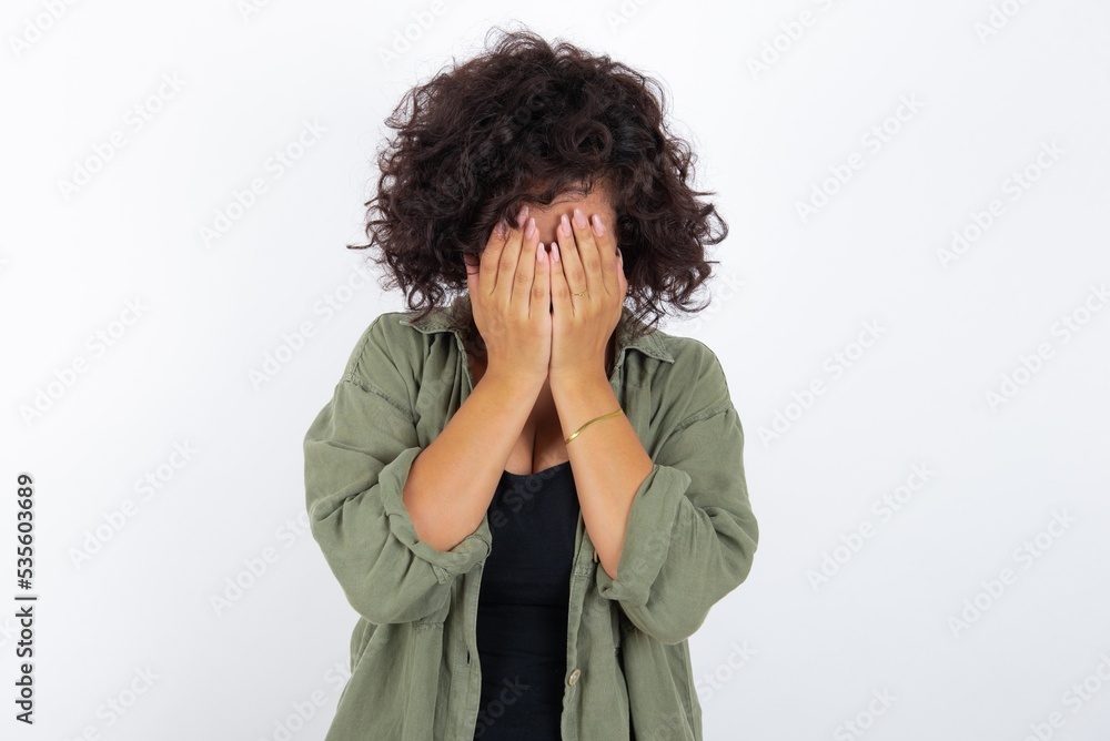 young beautiful woman with curly short hair wearing green overshirt over white wall covering her face with her hands, being devastated and crying. Sad concept young beautiful woman with curly short hair wearing green overshirt over white wall covering her face with her hands, being devastated and crying. Sad concept